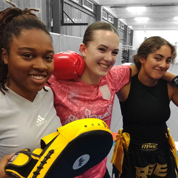 Trois femmes souriantes, se tenant les unes aux autres et aimant la pratique de la boxe thaïlandaise.